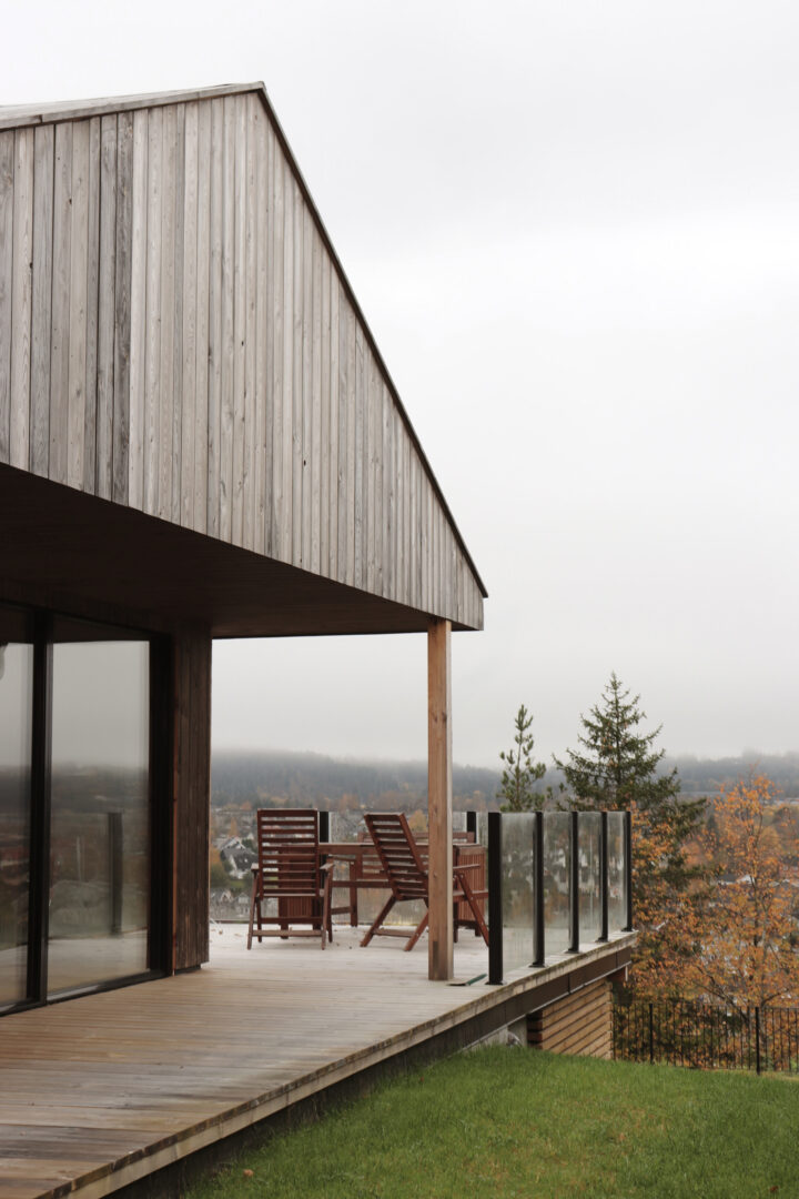 Wooden house exterior with a long deck, brown chairs, and glass railing overlooking a wooded valley on a cloudy day