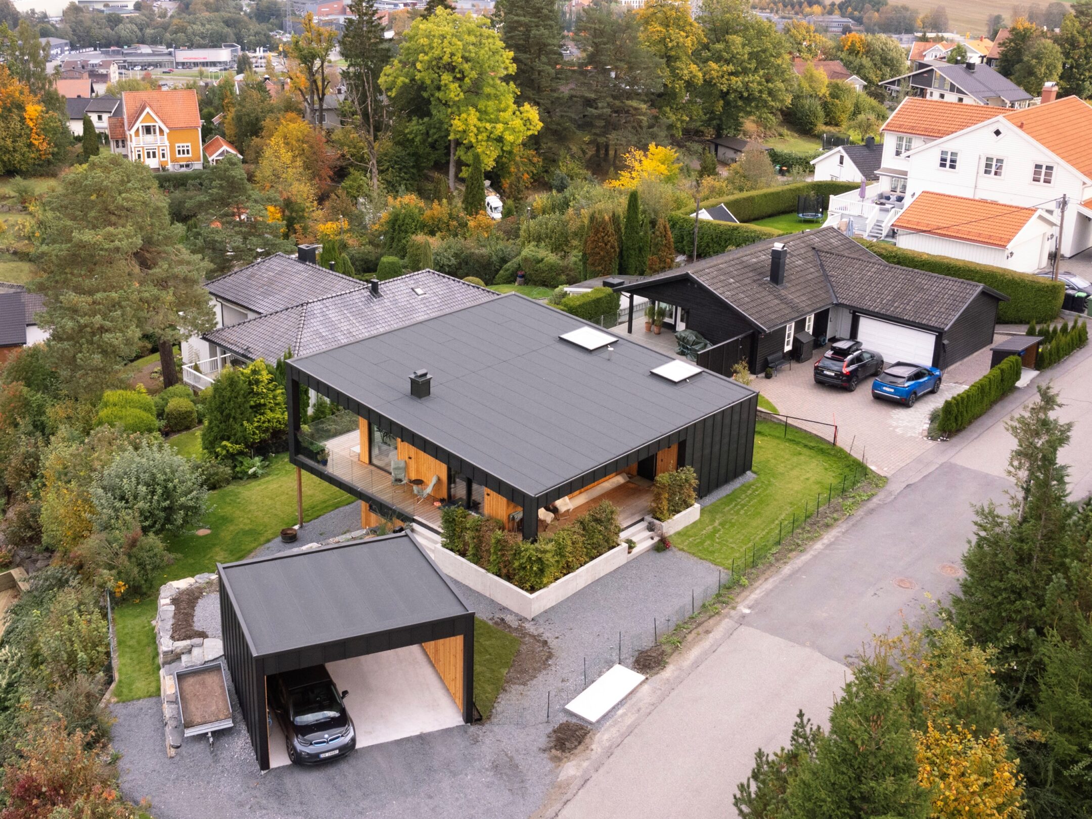 Aerial view of a modern black and wood house with a two-car garage, surrounding greenery and neighboring homes on a tree-lined street.