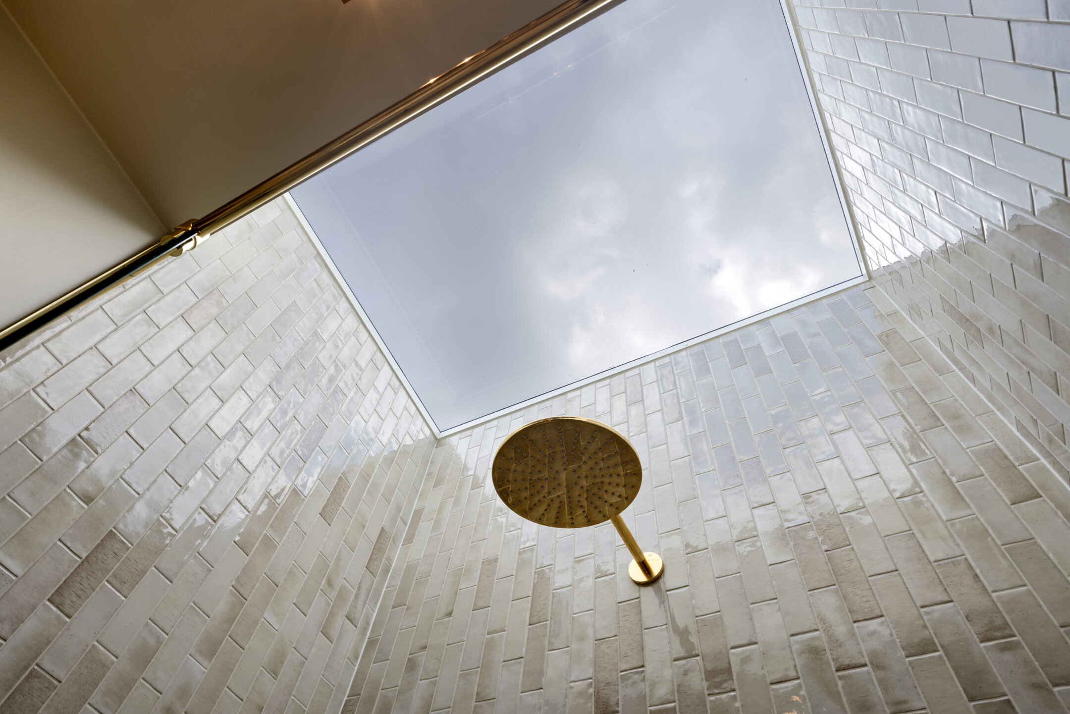 Shower with light beige rectangular tiles and a gold circular rainfall showerhead mounted on a vertical bar.