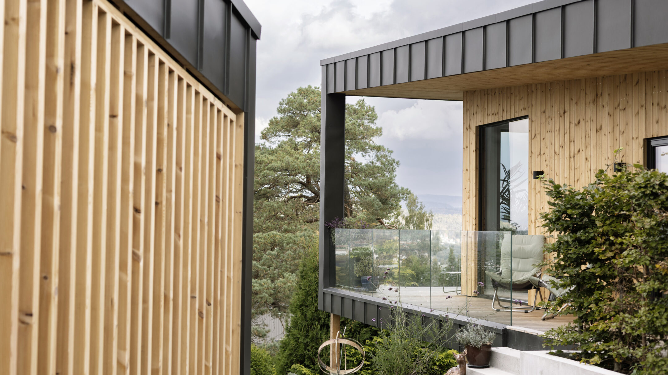Modern wooden house exterior with glass balcony and gray metal roof, surrounded by greenery and trees outside.