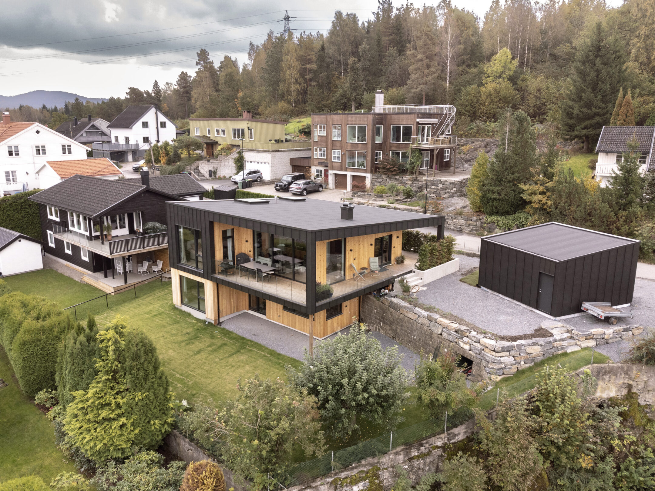 A modern glass-front house with wooden panels overlooks a green yard and neighboring homes in a hilly neighborhood at the edge of trees.