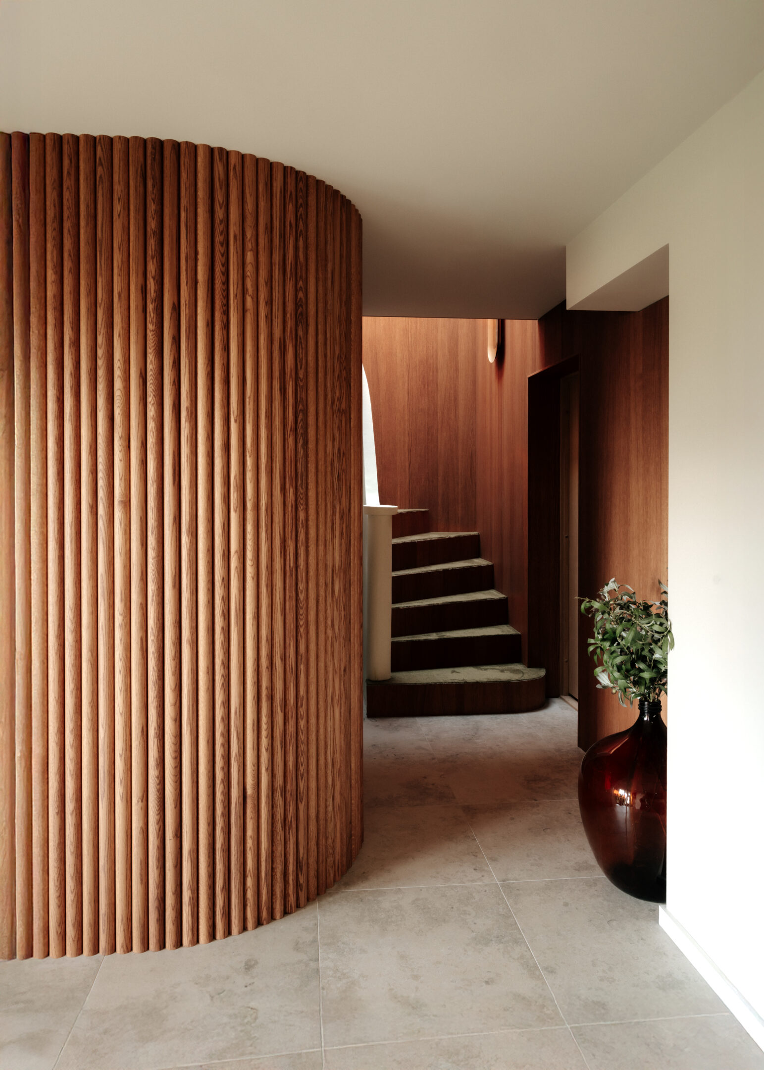 Curved vertical wooden slat wall in a modern hallway, with stairs in the background and a large brown vase with a plant to the right.