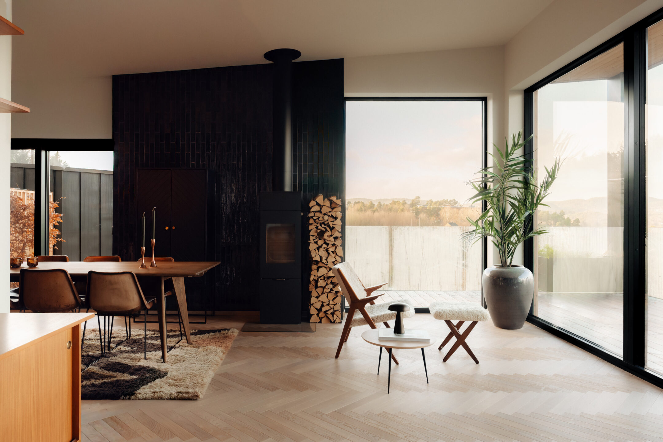 Open-plan living and dining area with a dark tiled fireplace, stacked firewood, and large floor-to-ceiling windows overlooking a terrace.