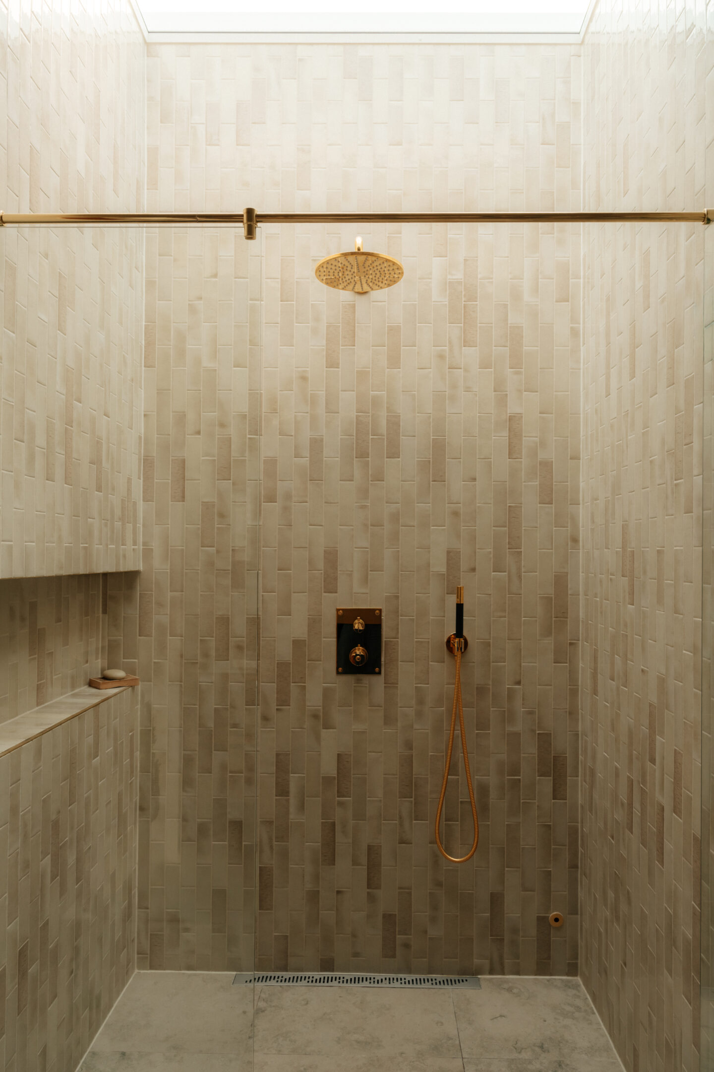 Beige tiled shower with a round gold rainfall showerhead and gold fixtures; handheld hose on the right, skylight above, recessed bench on the left.