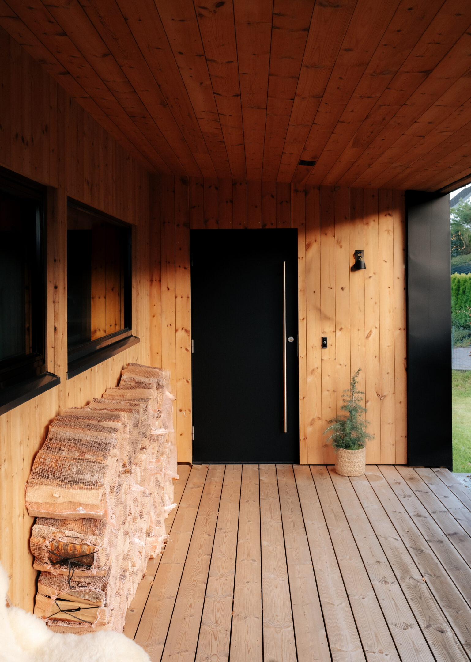Wood-clad porch with a black front door, stacked firewood wrapped in netting along the left wall, and a potted evergreen by the entrance.