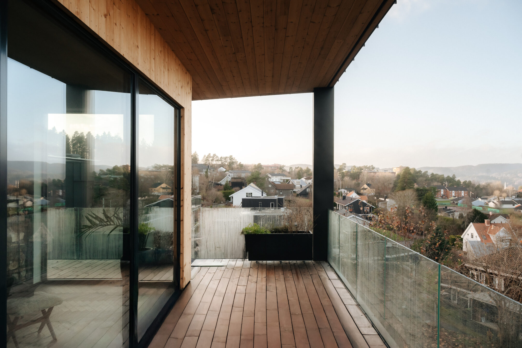 Wooden balcony with a glass railing, looking out over a suburban neighborhood on a clear day.